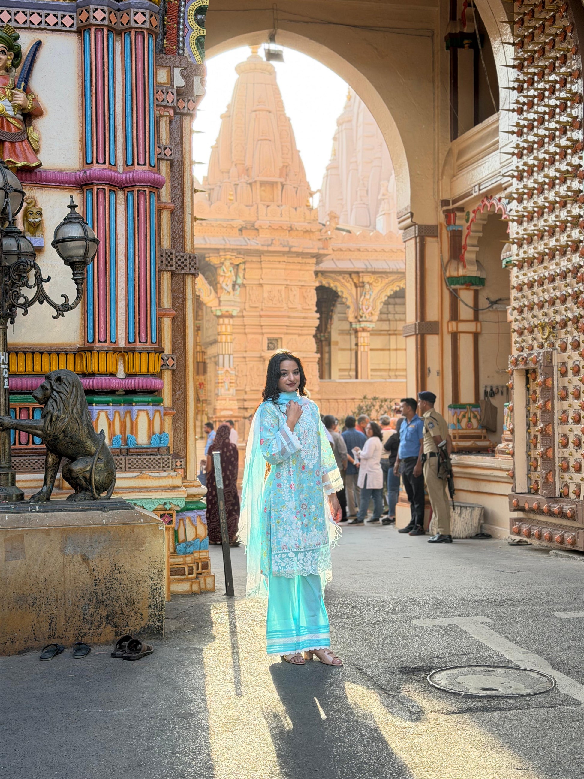 Woman in a blue traditional outfit standing in a temple courtyard with architectural details and people in the background.